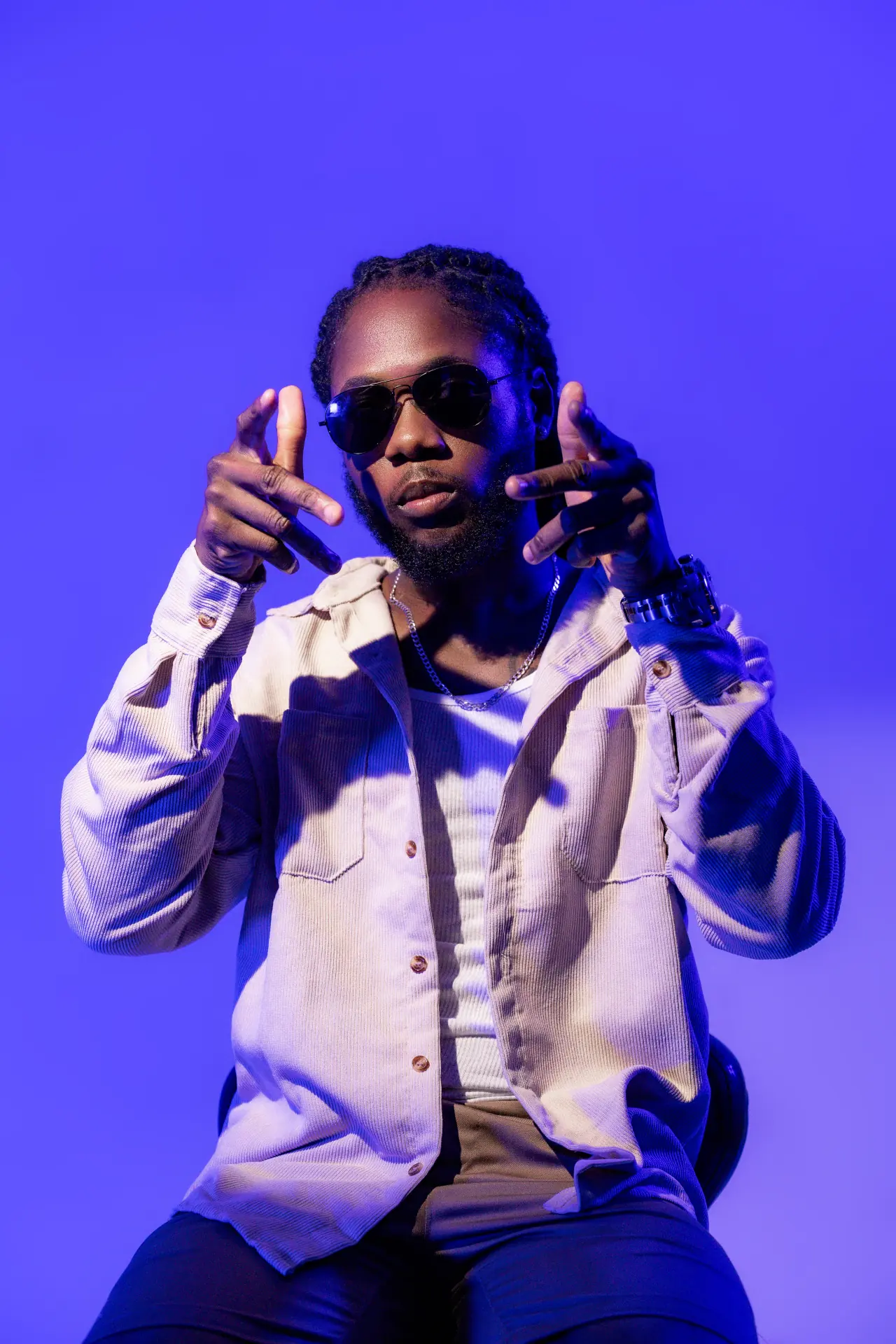 Saint Chris seated in blue light wearing sunglasses and cream overshirt.
